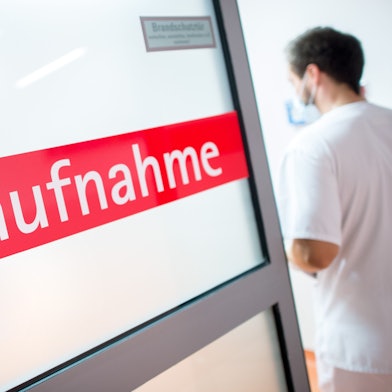 Ein Krankenpfleger steht in einem Krankenhaus neben einer Glastür mit der Aufschrift Notaufnahme.
