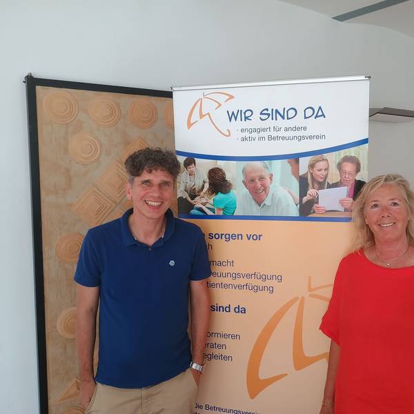 Frank Nixdorf steht links im Bild und trägt ein blaues Polo-Hemd. Rechts steht Anette Gövert, sie trägt ein rotes Kleid. Hinter den beiden ist ein Banner aufgestellt, auf dem die Aufschrift steht: „Wir sind da. Engagiert für andere, aktiv im Betreuungsverein.“