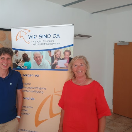 Frank Nixdorf steht links im Bild und trägt ein blaues Polo-Hemd. Rechts steht Anette Gövert, sie trägt ein rotes Kleid. Hinter den beiden ist ein Banner aufgestellt, auf dem die Aufschrift steht: „Wir sind da. Engagiert für andere, aktiv im Betreuungsverein.“