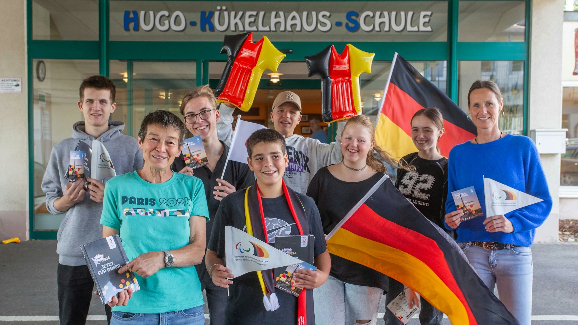 Mehrere Schüler und zwei Betreuerinnen stehen mit Deutschlandflaggen und weiteren Fan-Artikel in Deutschland-Farben vor einer Schule.