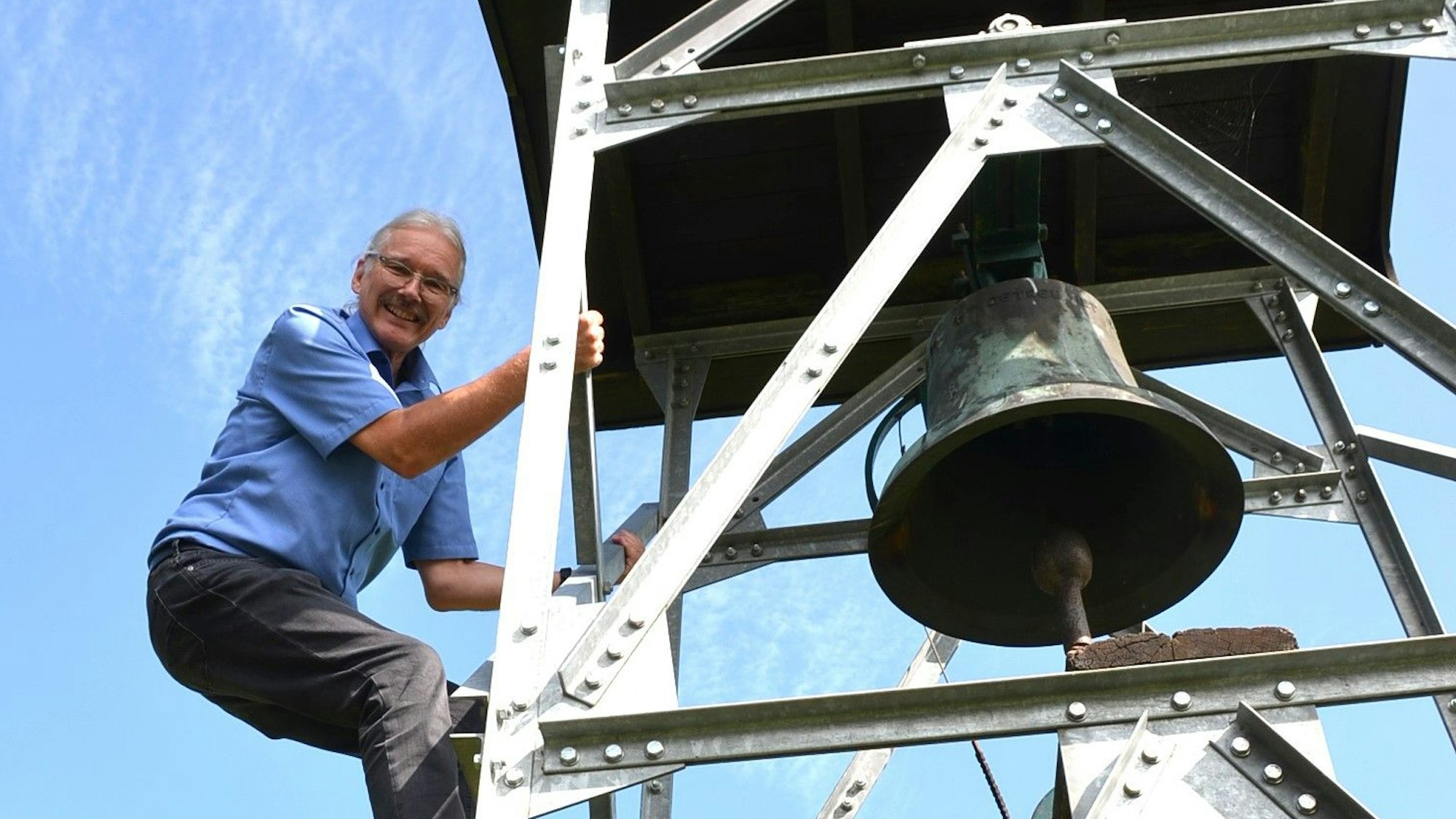 Der kleine Friedhof in der Waldbröler Ortschaft Seifen hat jetzt einen Glockenturm. Dieser stammt aus dem Nachbarort Helten. Pfarrer Thomas Seibel klettert hinauf, um die Inschrift auf der Glocke zu entziffern.