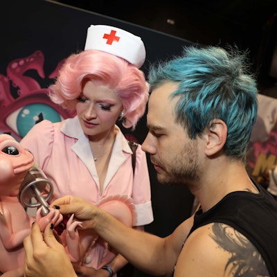 Eine Frau im rosafarbenen Krankenschwester Kostüm hält eine Plastikfigur im Arm, an der ein Mann mit blauen Haaren noch etwas richtet.