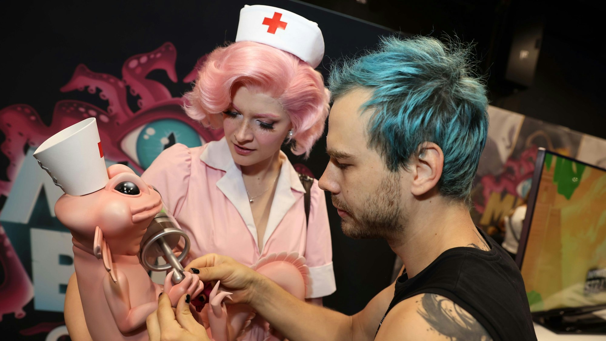 Eine Frau im rosafarbenen Krankenschwester Kostüm hält eine Plastikfigur im Arm, an der ein Mann mit blauen Haaren noch etwas richtet.