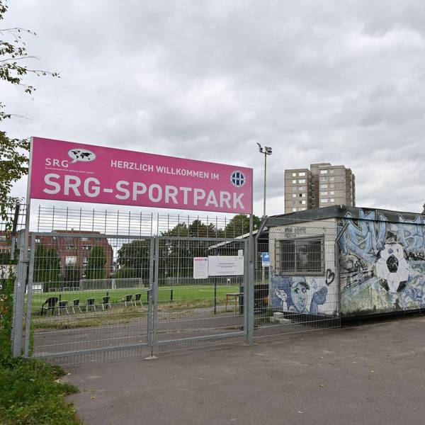 Im Vordergrund ist der abgesperrte Eingang zum Fußballplatz. Im Hintergrund sieht man die Hochhäuser.