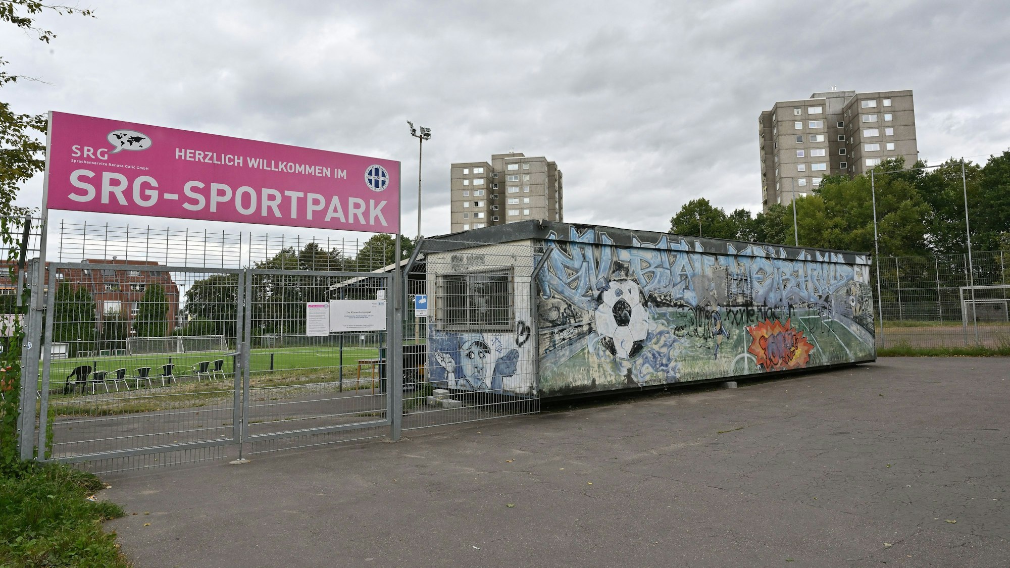 Im Vordergrund ist der abgesperrte Eingang zum Fußballplatz. Im Hintergrund sieht man die Hochhäuser.
