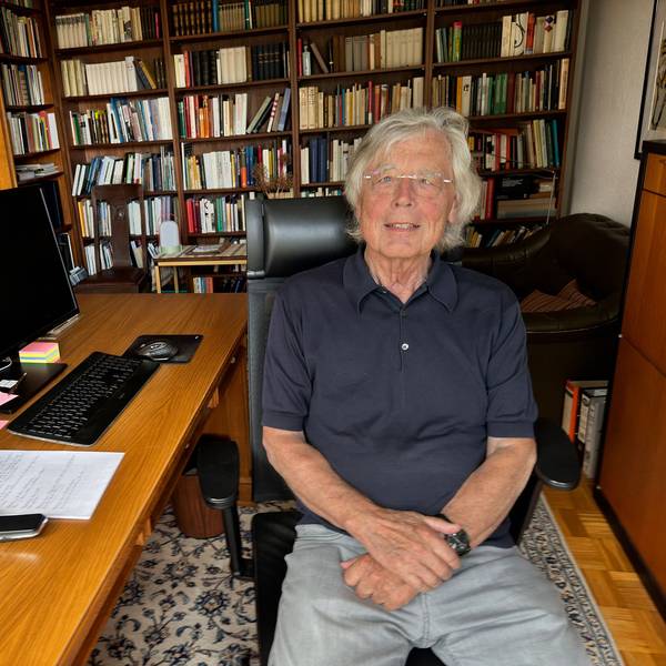 Auf dem Bild ist Wißkirchen zu sehen. Er sitzt auf ienem Bürostuhl am Schreibtisch und trägt ein dunkelblaues Polohemd und eine graue Hose. Im Hintergrund das Arbeitszimmer, an den Wänden stehen volle Bücherschränke.