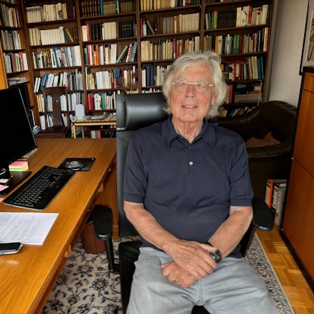 Auf dem Bild ist Wißkirchen zu sehen. Er sitzt auf ienem Bürostuhl am Schreibtisch und trägt ein dunkelblaues Polohemd und eine graue Hose. Im Hintergrund das Arbeitszimmer, an den Wänden stehen volle Bücherschränke.