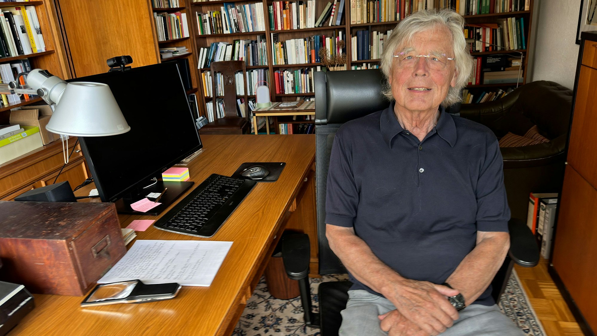 Auf dem Bild ist Wißkirchen zu sehen. Er sitzt auf ienem Bürostuhl am Schreibtisch und trägt ein dunkelblaues Polohemd und eine graue Hose. Im Hintergrund das Arbeitszimmer, an den Wänden stehen volle Bücherschränke.