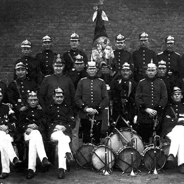 Feuerwehrleute in Drillichröcken und mit Musikkappelle stehen um 1900 zu einem Gruppenbild zusammen.