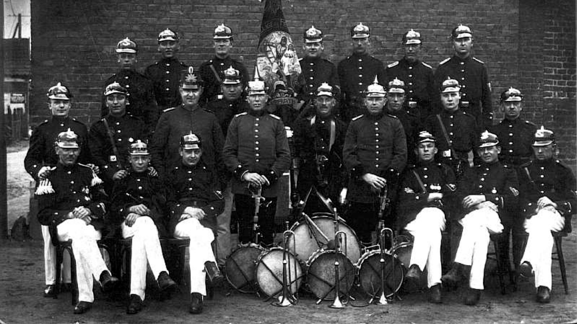 Feuerwehrleute in Drillichröcken und mit Musikkappelle stehen um 1900 zu einem Gruppenbild zusammen.