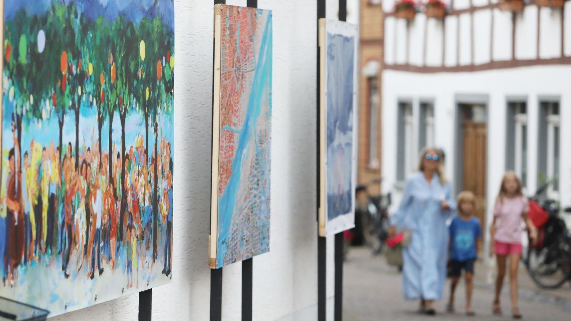 Drei Gemälde nebeneinander, im Hintergrund eine Frau im blauen Kleid mit zwei Kindern auf der Straße.