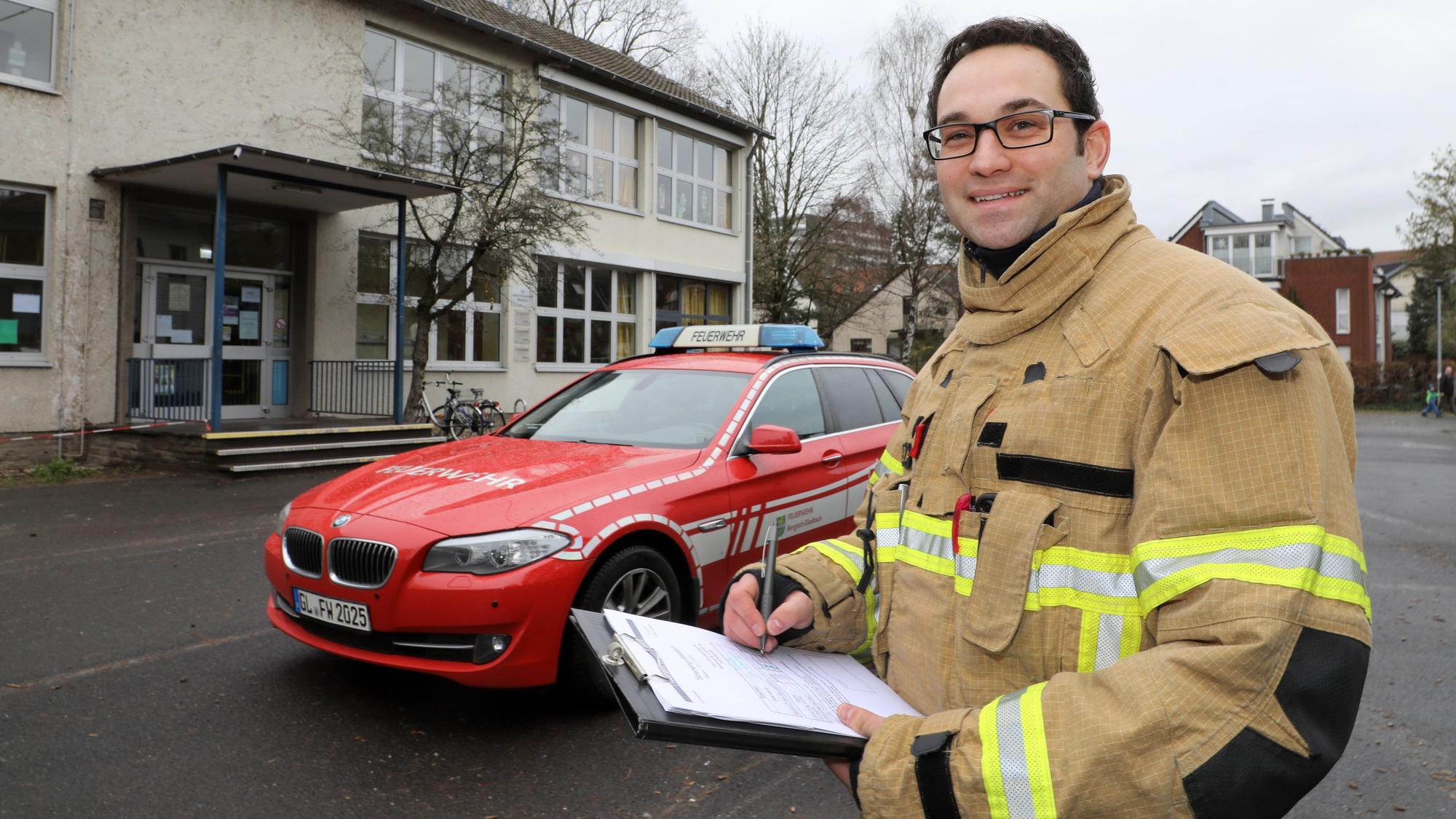 Löschzugführer Felix Vorndran steht mit einem Klemmbrett vor einem Feuerwehrauto auf einem Refrather Schulhof.