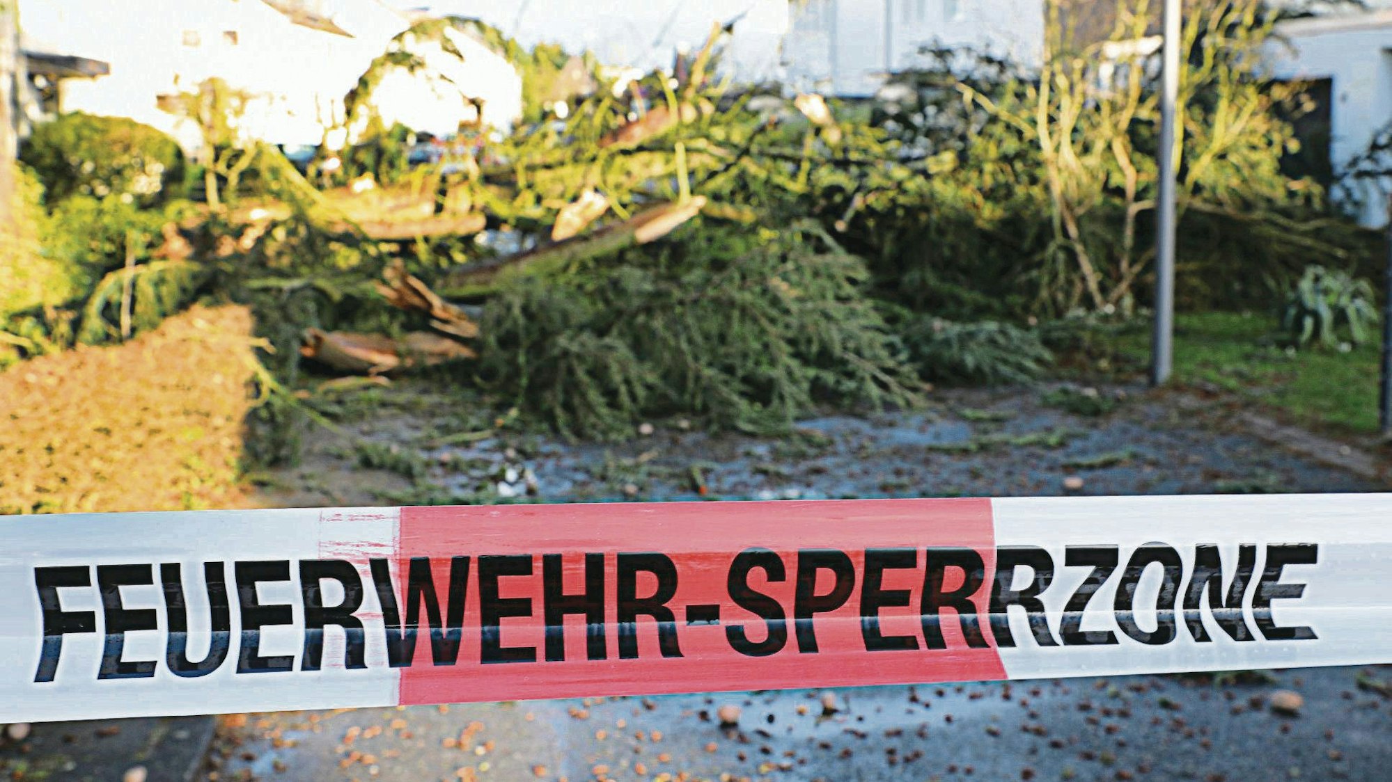 Ein rot-weißes Flatterband mit der Aufschrift „Feuerwehr-Sperrzone“ sperrt eine Straße, über die Bäume gestürzt sind.