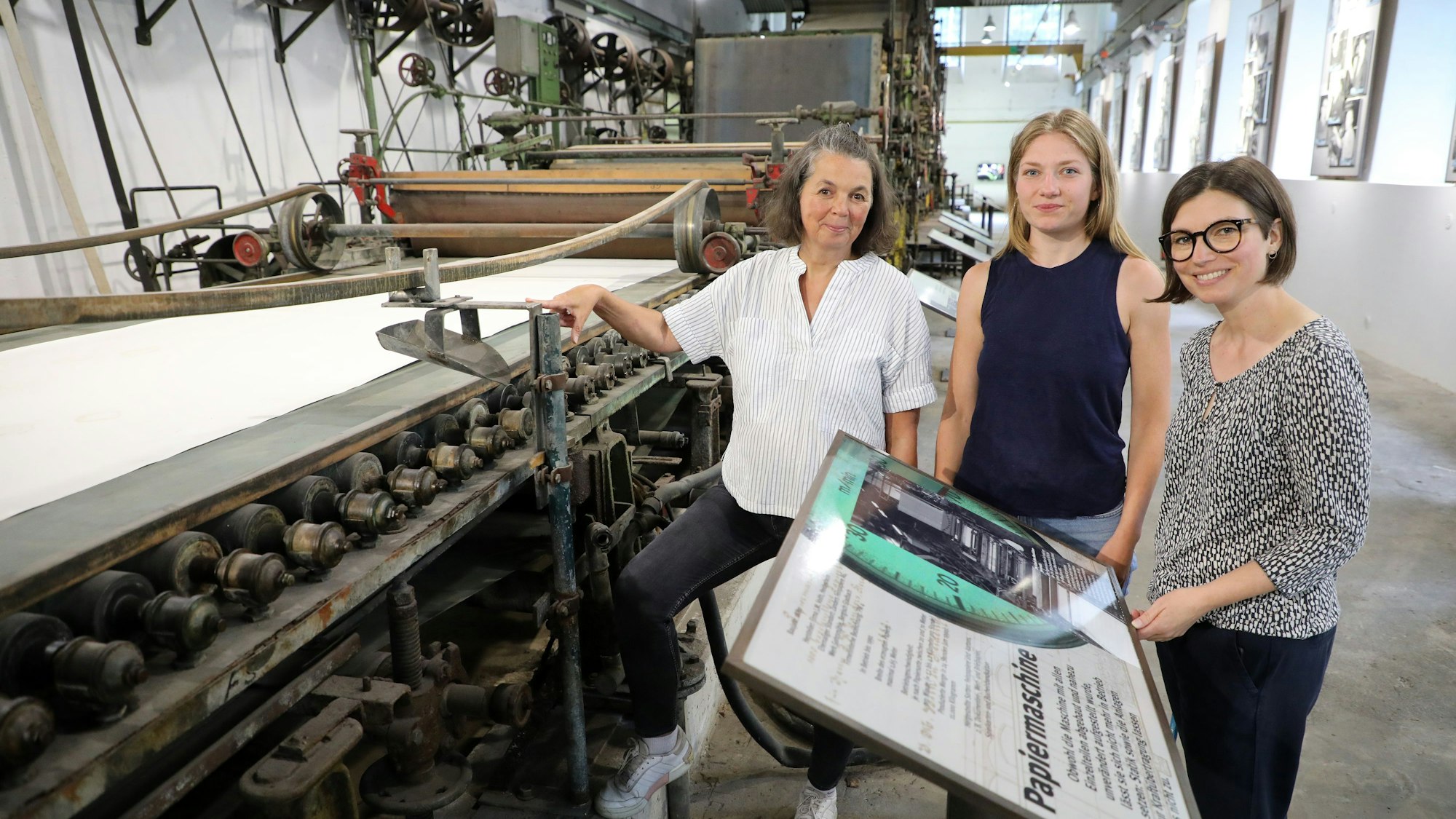 Drei Frauen stehen an der Papiermaschine 4 des LVR-Industriemuseums Papiermühle Alte Dombach in Bergisch Gladbach.