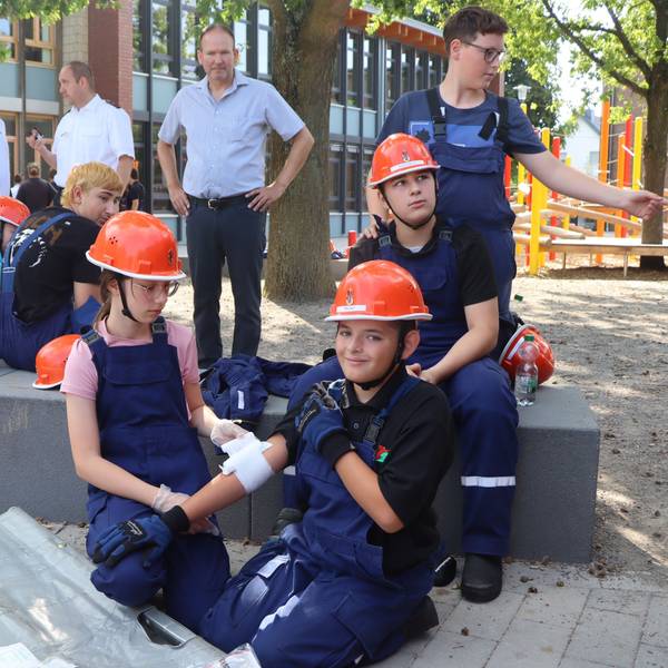 Auf dem Bild legt ein Mädchen eine Bandage um den Arm eines Jungen. Mehrere Kinder schauen zu. Sie alle tragen blaue Overalls und orangene Helme.