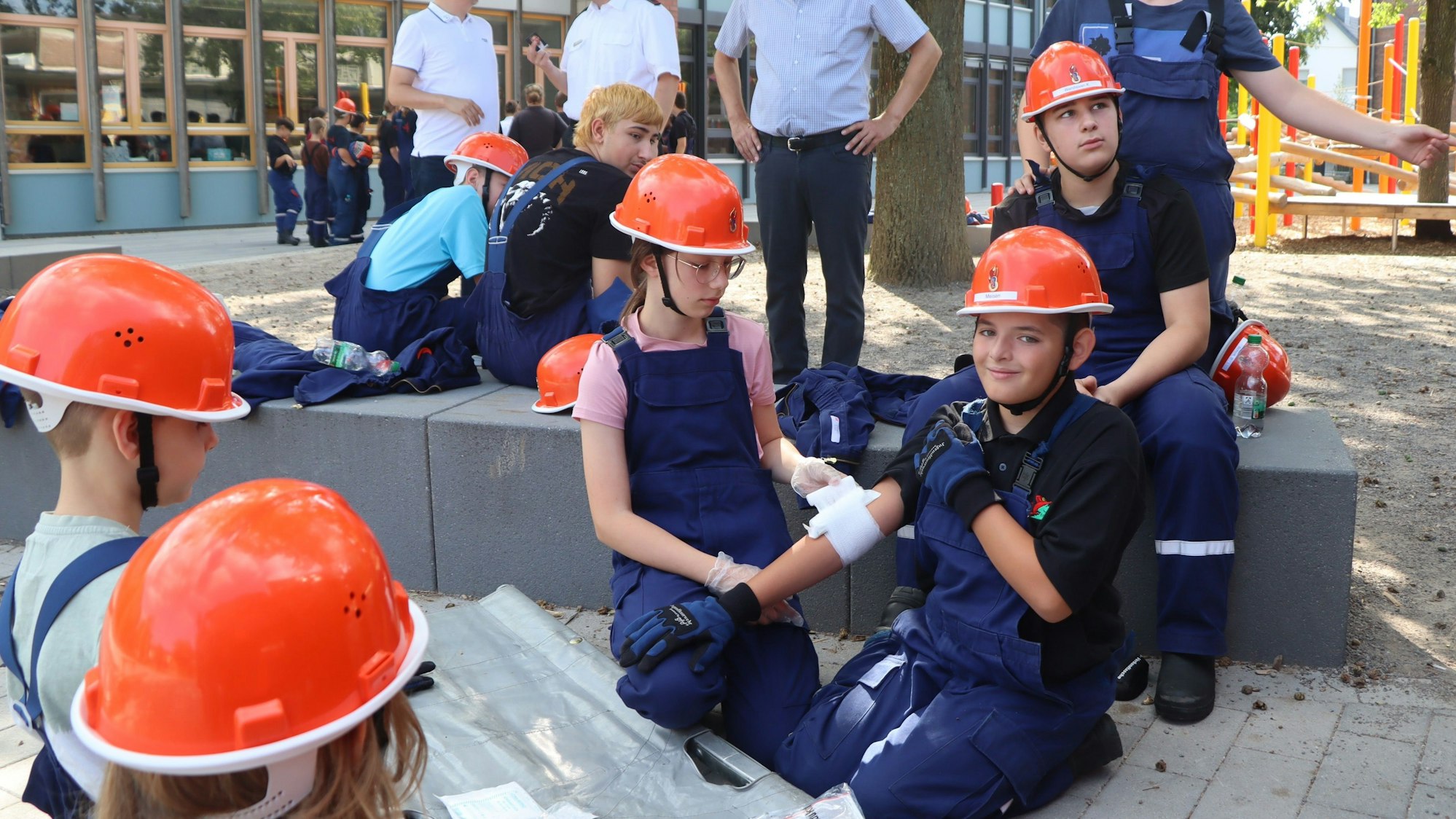 Auf dem Bild legt ein Mädchen eine Bandage um den Arm eines Jungen. Mehrere Kinder schauen zu. Sie alle tragen blaue Overalls und orangene Helme.