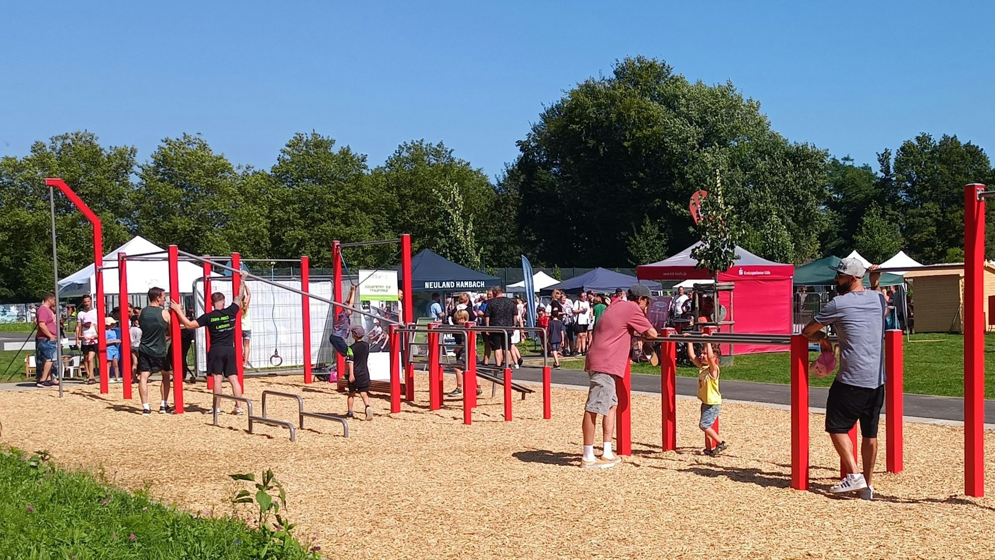 Auf dem Bild ist ein Sportareal zu sehen, auf dem verschiedene rote Stangen als Turn- und Fitnessgeräte dienen. Einige Menschen probieren die Gerätschaften aus.