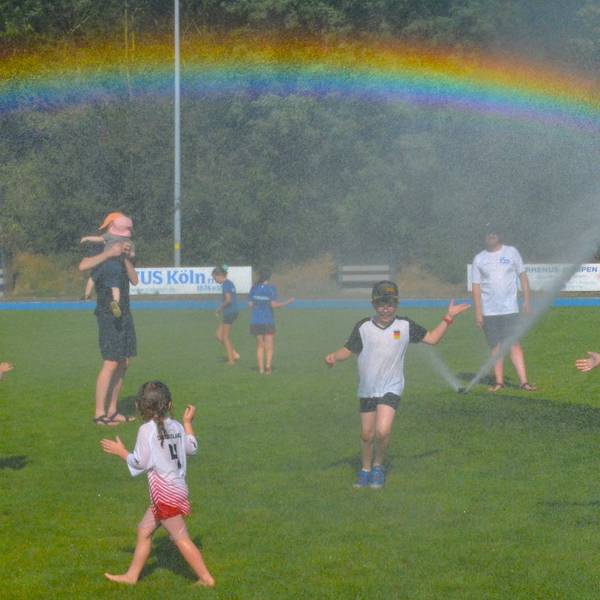 Kinder laufen über eine Wiese. Ein Rasensprenger sorgt für kühles Nass und einen Regenbogen.