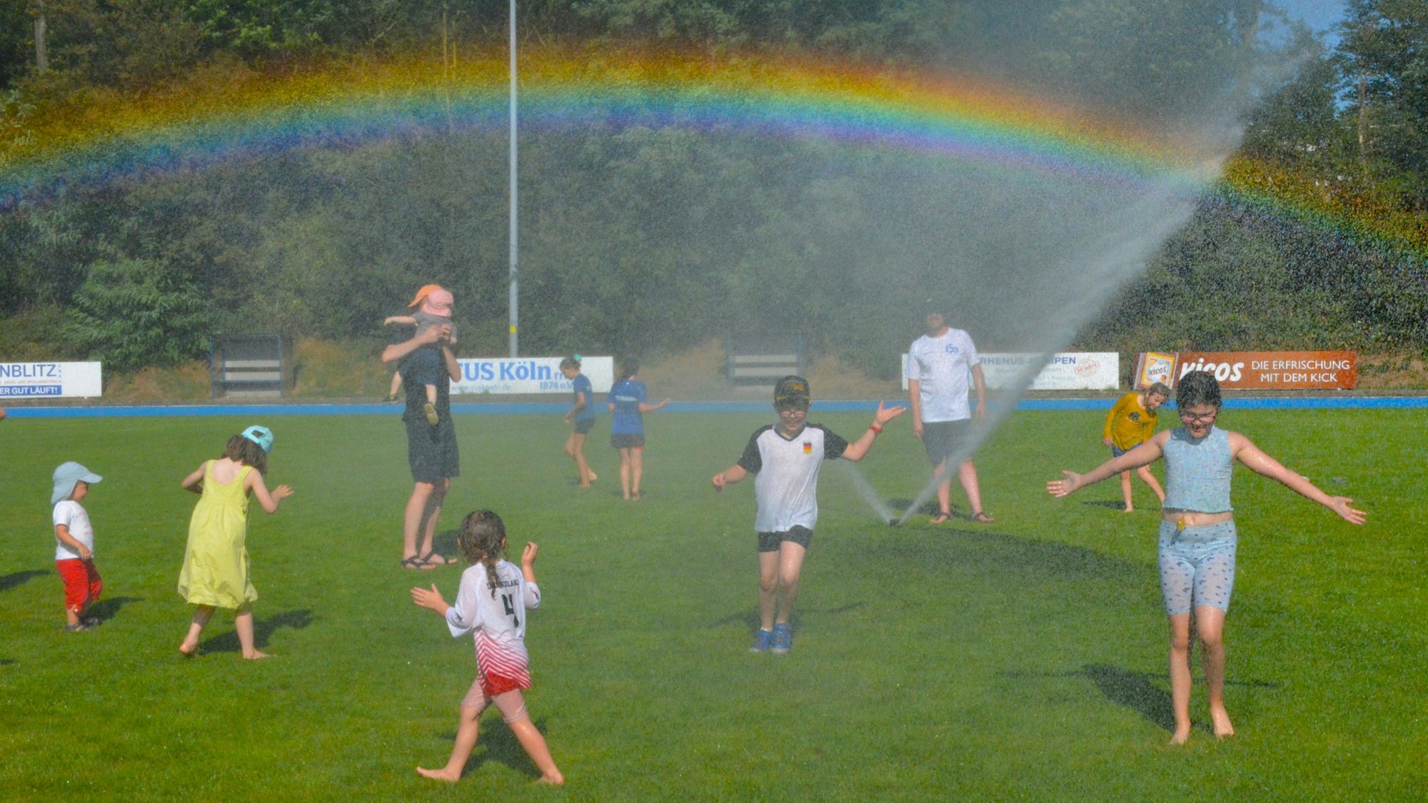 Kinder laufen über eine Wiese. Ein Rasensprenger sorgt für kühles Nass und einen Regenbogen.