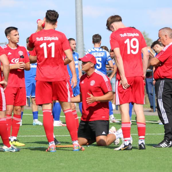 Zülpichs Trainer kniet während einer Trinkpause inmitten seiner Spieler auf dem Rasen.
