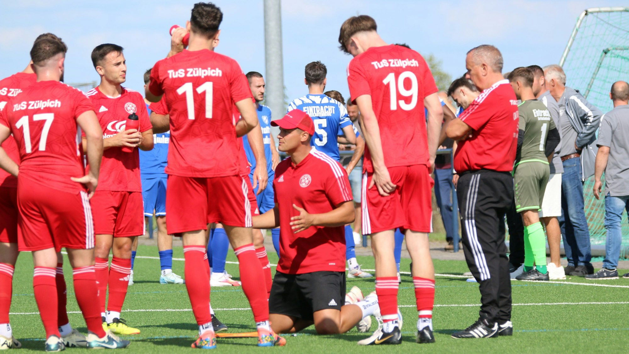 Zülpichs Trainer David Sasse kniet auf dem Spielfeld. Seine Spieler stehen im Halbkreis um ihn herum und hören zu.