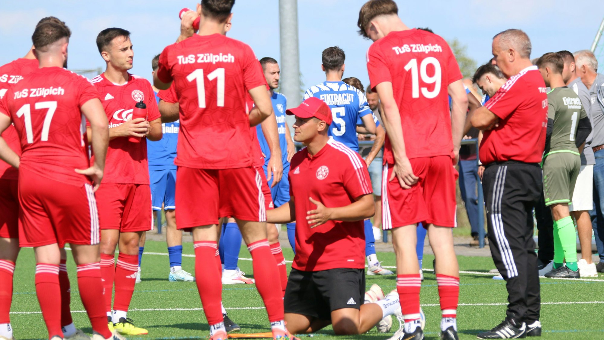 Zülpichs Trainer kniet während einer Trinkpause inmitten seiner Spieler auf dem Rasen.
