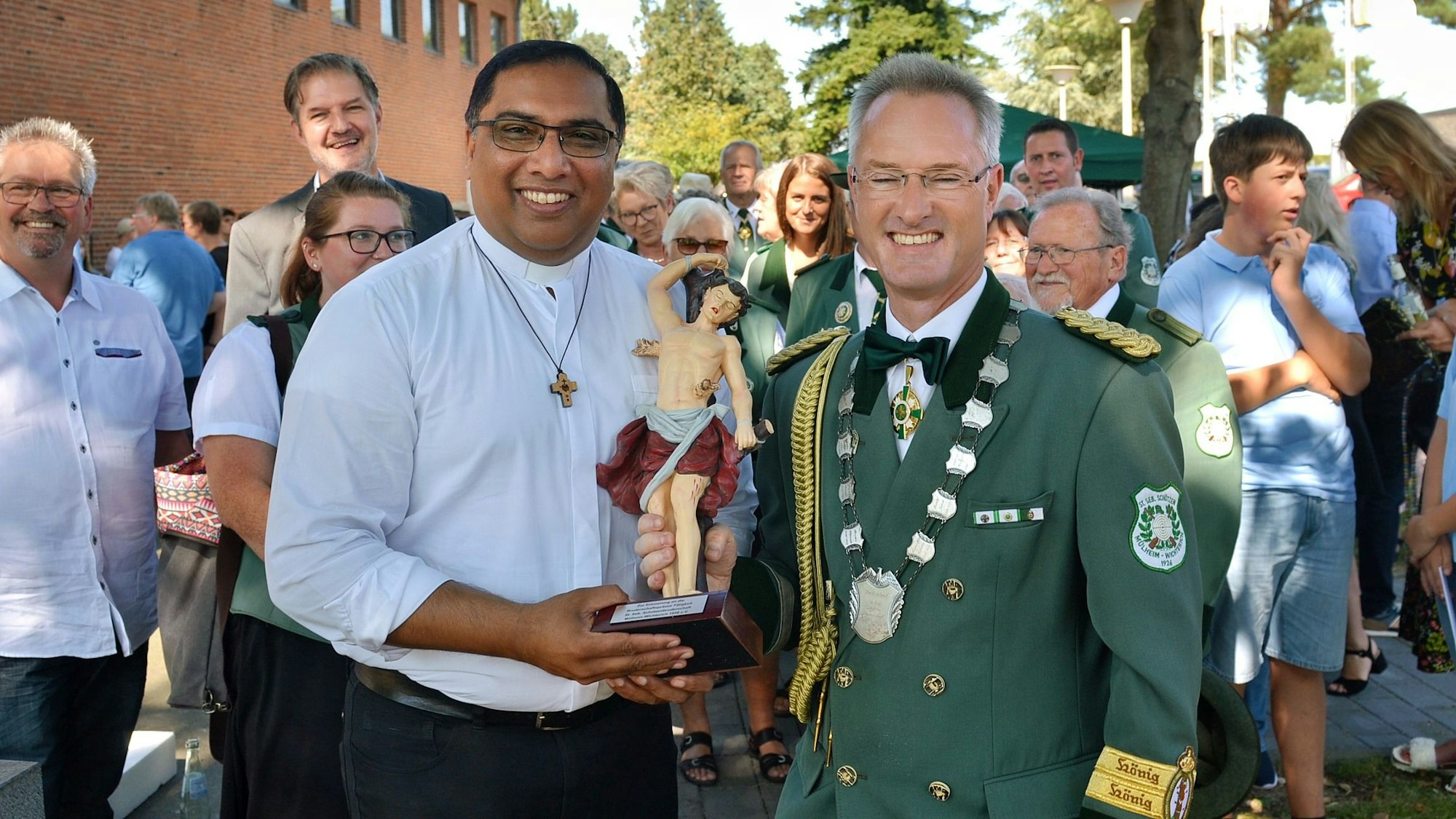 Ein Mann mit weißem Hemd und Kreuzkette erhält eine Figur von einem Mann in grüner Schützen-Uniform.