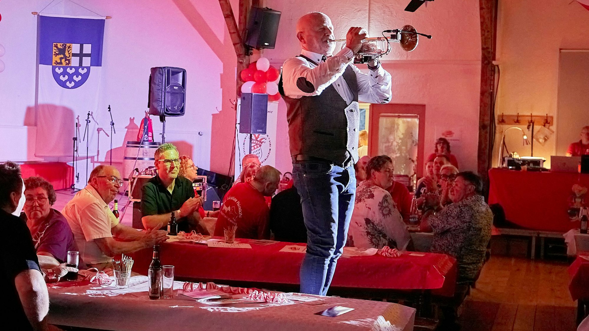 Ein Mann steht Trompete spielend auf einer Bierbank in einem rot ausgeleuchteten Saal, um ihn herum sitzt Publikum.
