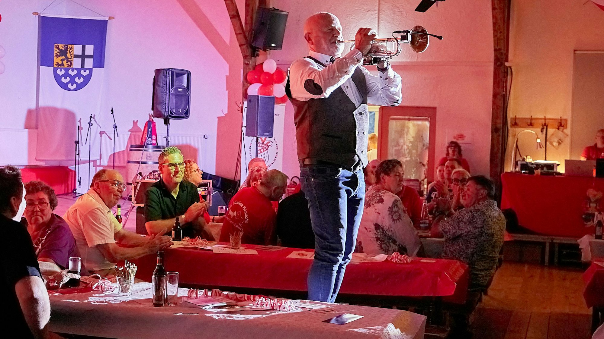 Ein Mann steht Trompete spielend auf einer Bierbank in einem rot ausgeleuchteten Saal, um ihn herum sitzt Publikum.