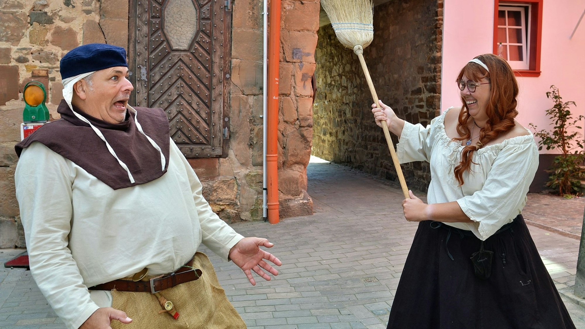 Eine mittelalterlich gewandete Frau erhebt lachend einen Besen gegen einen mittelalterlich gewandeten Mann, der eine erschreckte Miene aufsetzt.