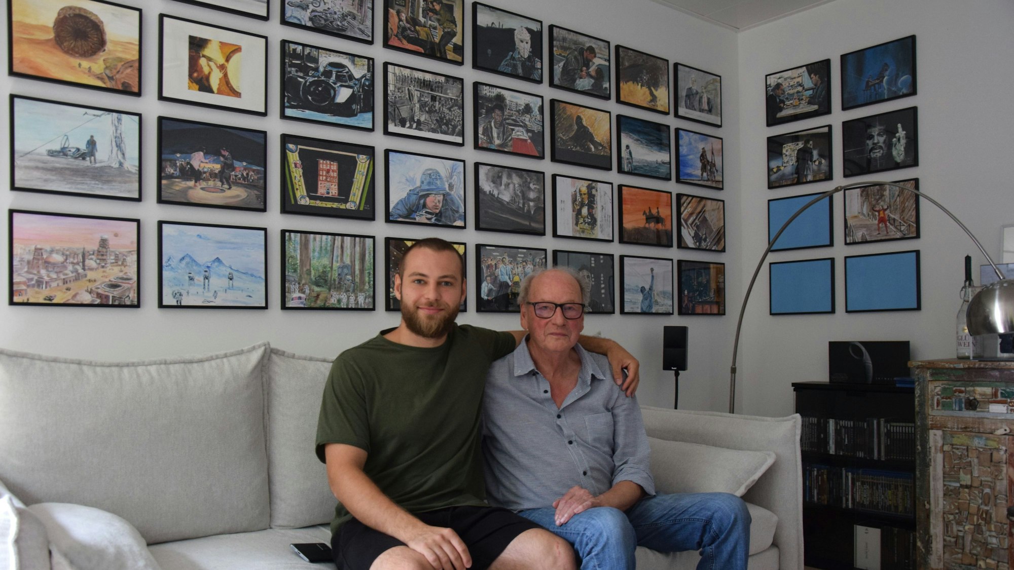 Zu sehen sind Winfried Brück (rechts) und sein Enkel. Sie sitzen auf einer Couch, im Hintergrund hängen eingerahmt die Filmszenen, die Brück für seinen Enkel gemalt hat.