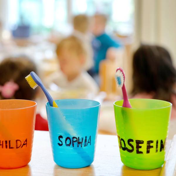 Zahnbürsten und Zahnputzbecher mit den Vornamen der Kinder stehen in einer Kindertagesstätte auf einem Tisch.