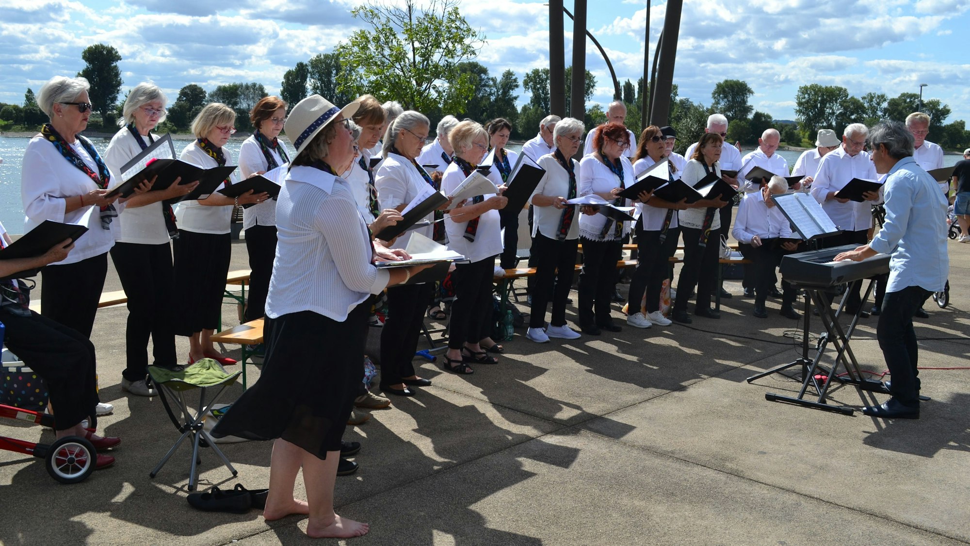 Männer und Frauen singen an einem Flussufer. Der Dirigent spielt an einem Keyboard.