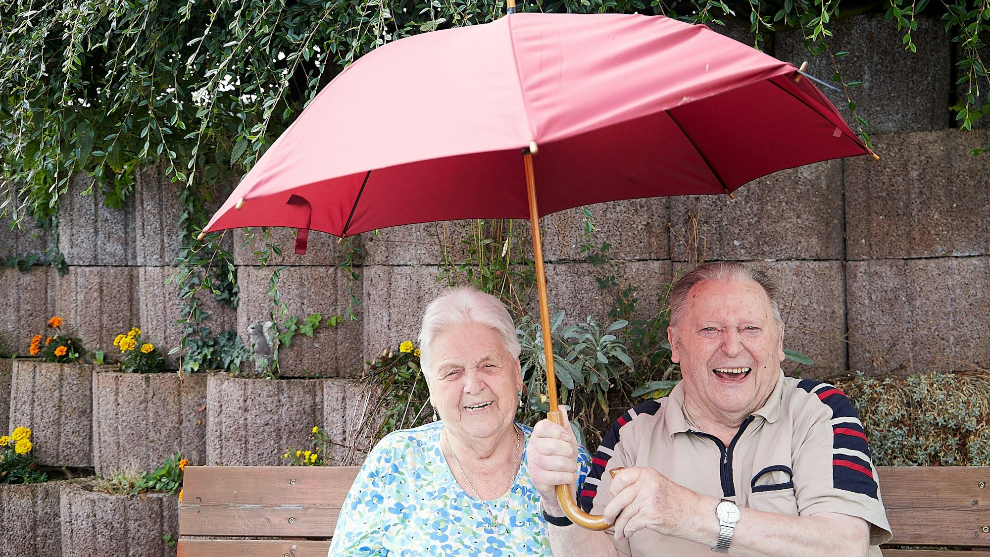 Eine alte Frau und ein alter Mann sitzen fröhlich auf einer Bank, während er einen Regenschirm hält.