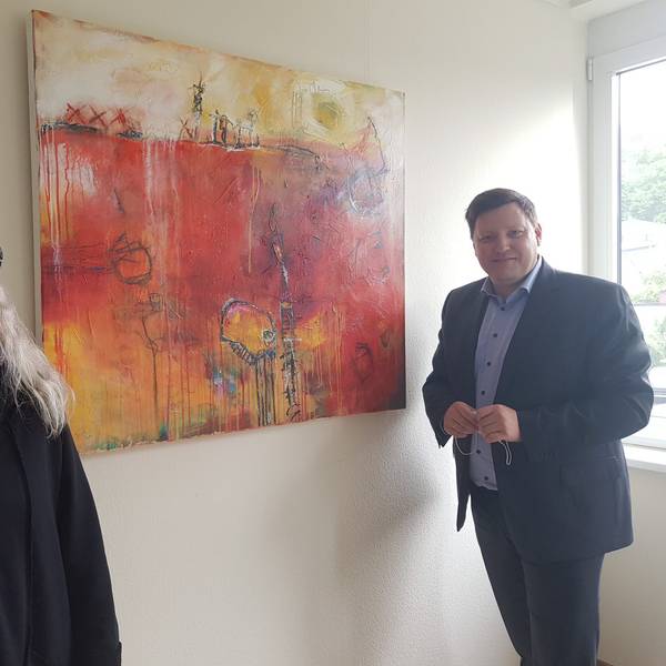 Ein Mann im blauen Sakko und Hemd neben einem abstrakten Gemälde in Rot- und Orangetönen.