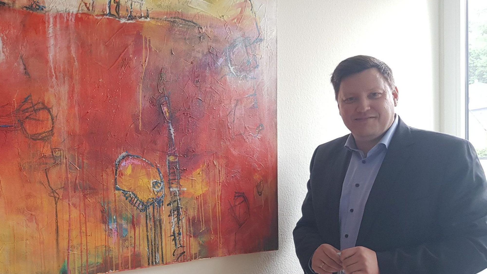 Ein Mann im blauen Sakko und Hemd neben einem abstrakten Gemälde in Rot- und Orangetönen.