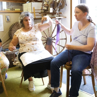 Die drei Frauen sitzen nebeneinander. Doris Schmitten bedient ein Spinnrad, ihre Mitstreiterinnen halten Fäden in den Händen.