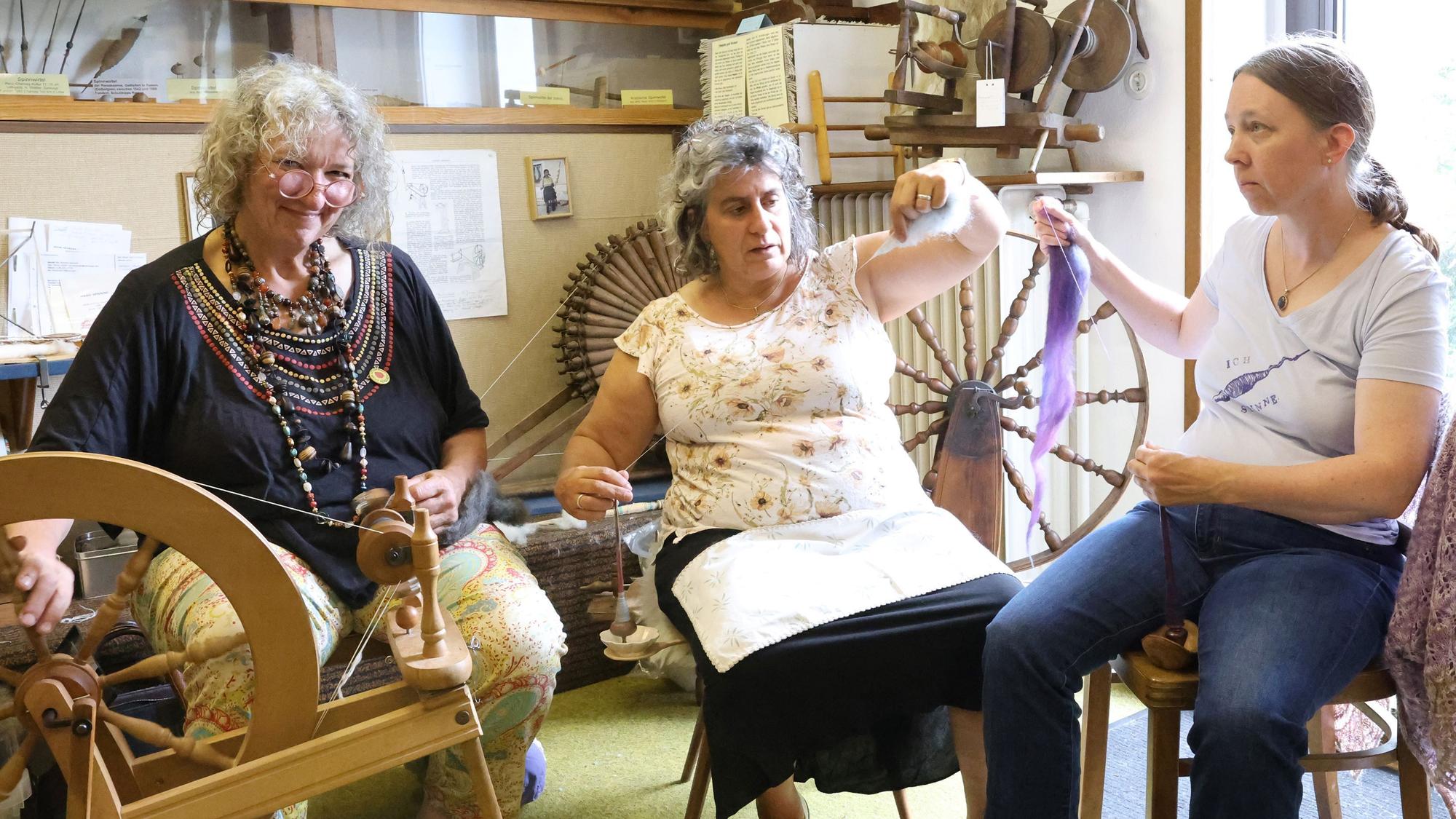 Die drei Frauen sitzen nebeneinander. Doris Schmitten bedient ein Spinnrad, ihre Mitstreiterinnen halten Fäden in den Händen.