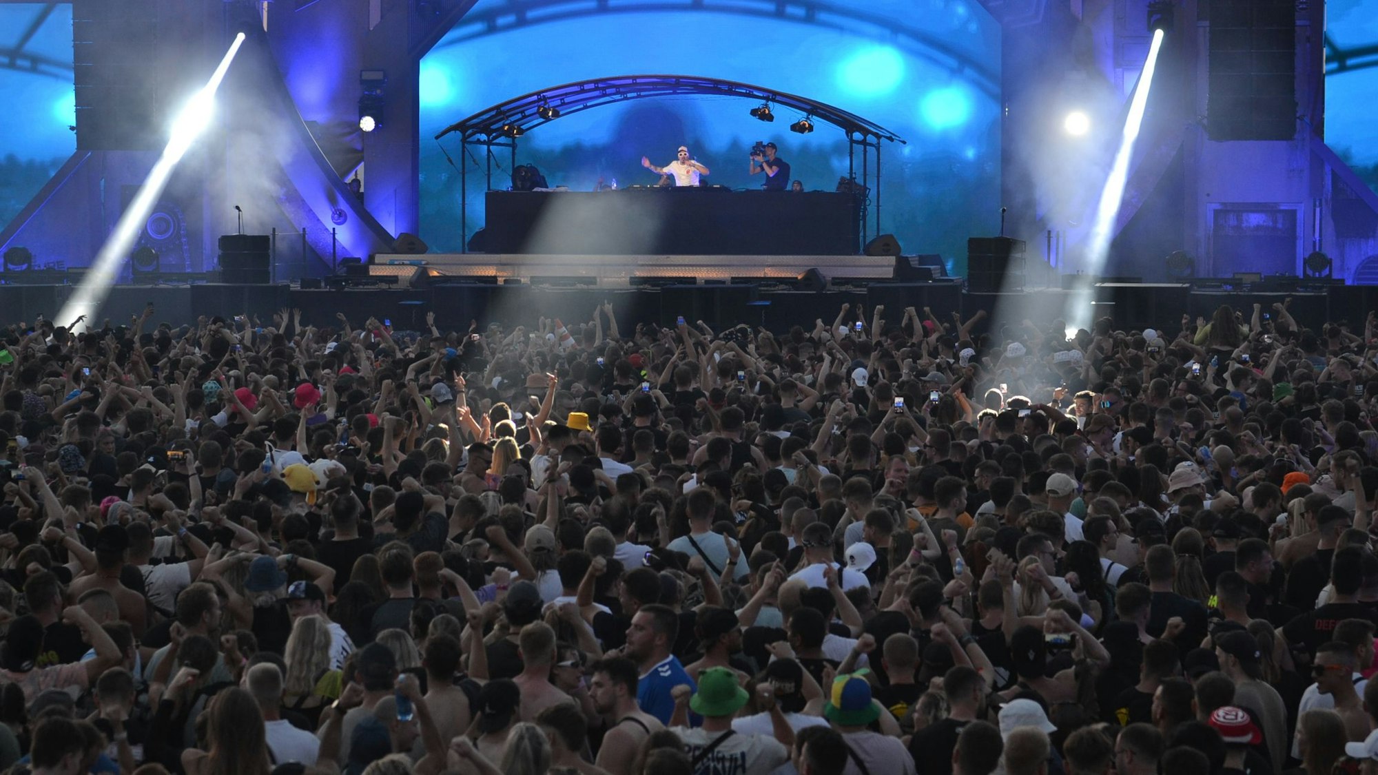 Sehr viele Menschen stehen vor einer mit hohen Elementen verkleideten Bühne, die in blaues Licht getaucht ist.