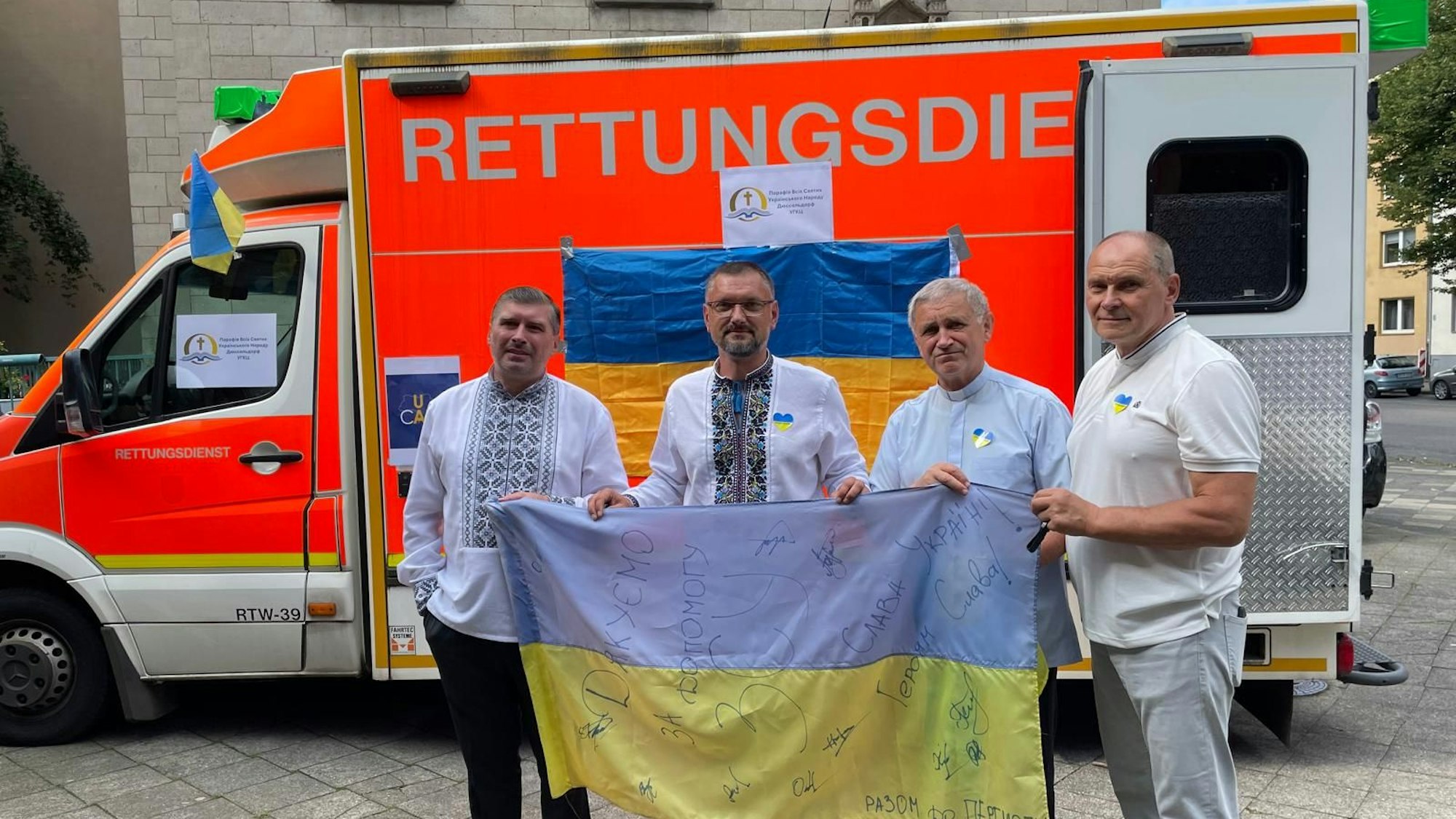 Vier Männer stehen mit einer ukrainischen Fahne in Duisburg vor einem Rettungswagen.