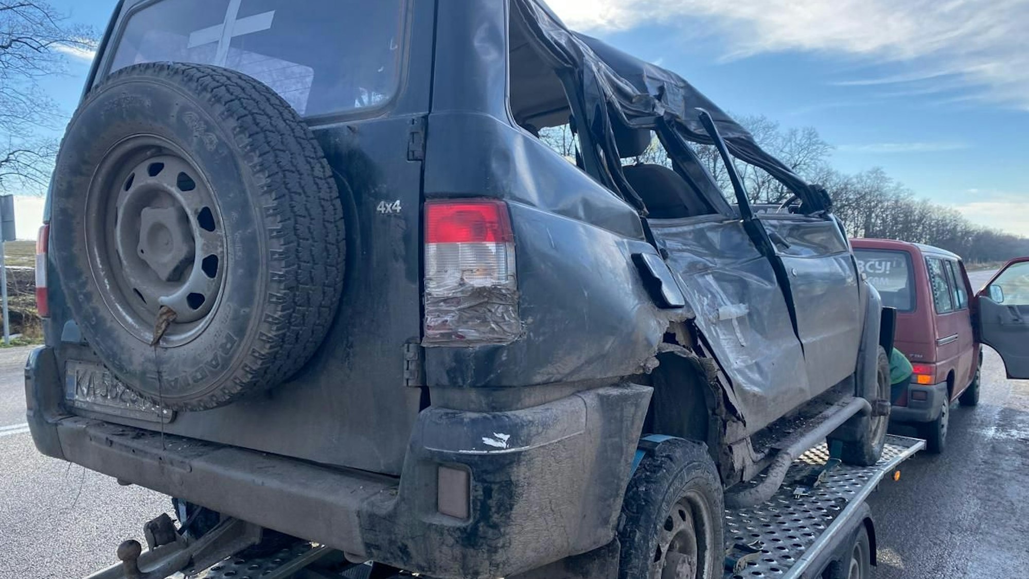 Ein stark demolierter Wagen steht auf einem Anhänger hinter einem roten VW-Bus.