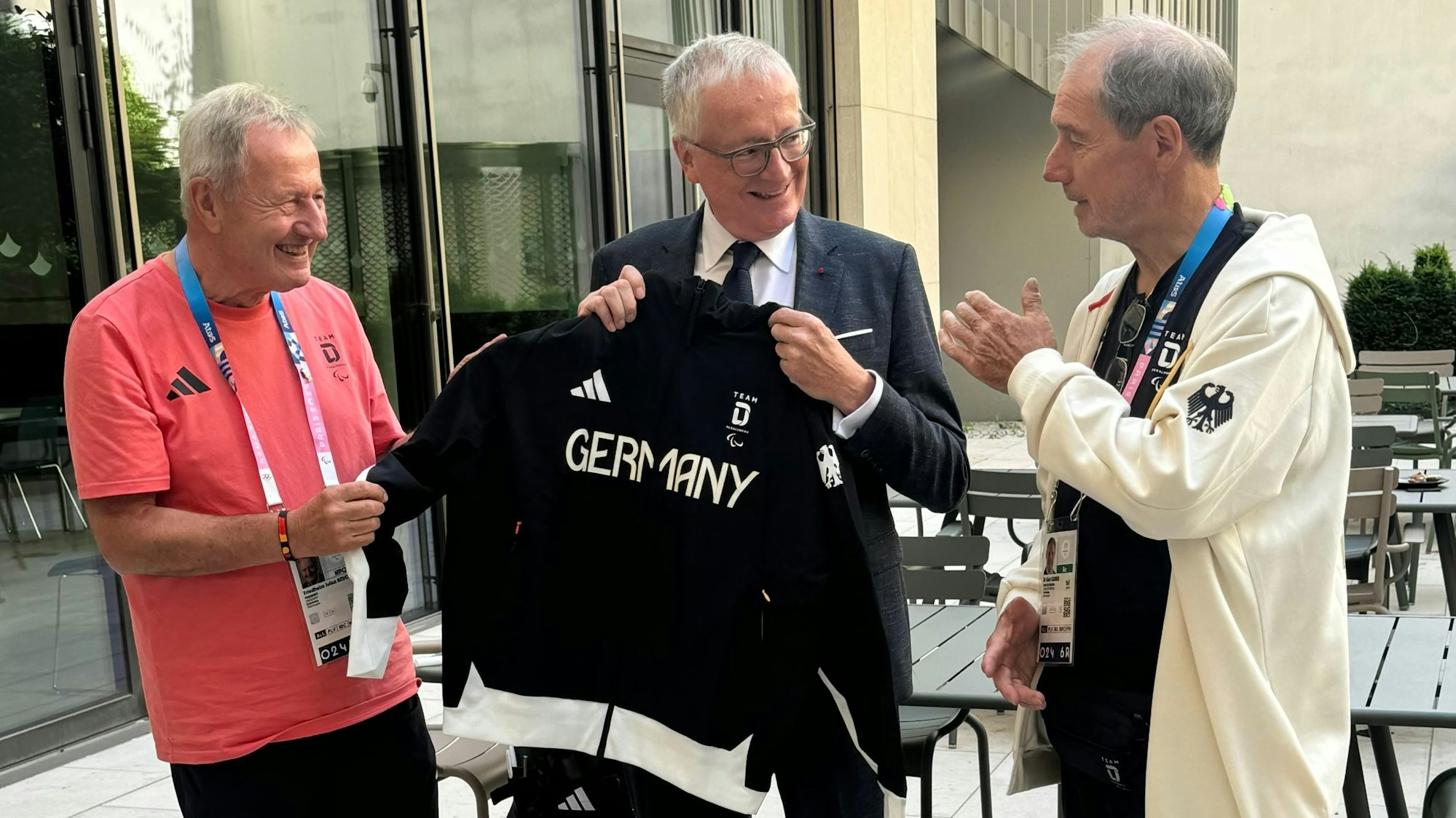 Drei Männer stehen beisammen. Einer hält einen schwarzen Trainingsanzug in der Hand, auf dem „Germany“ steht.
