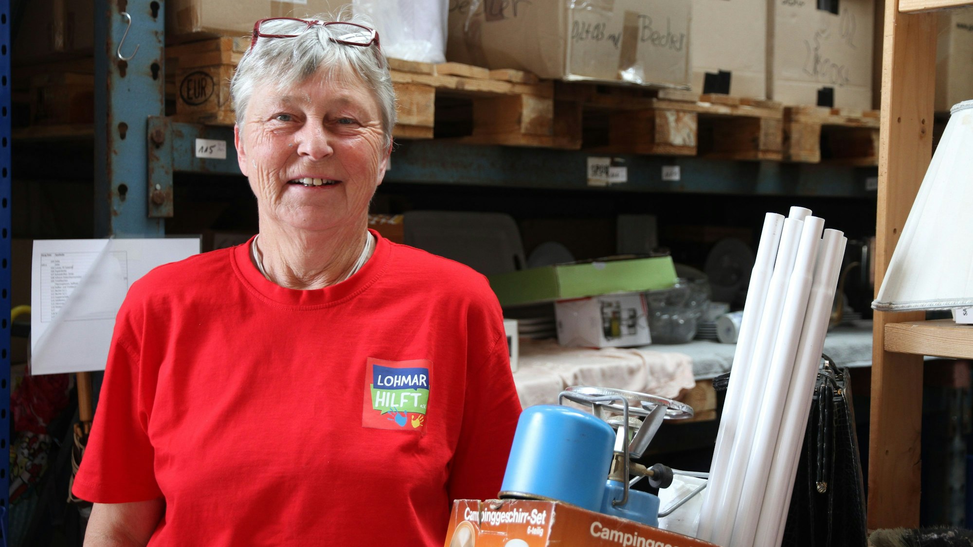 Eine Frau mit kurzen grauen Haaren und einem roten T-Shirt steht in einer Lagerhalle.ü