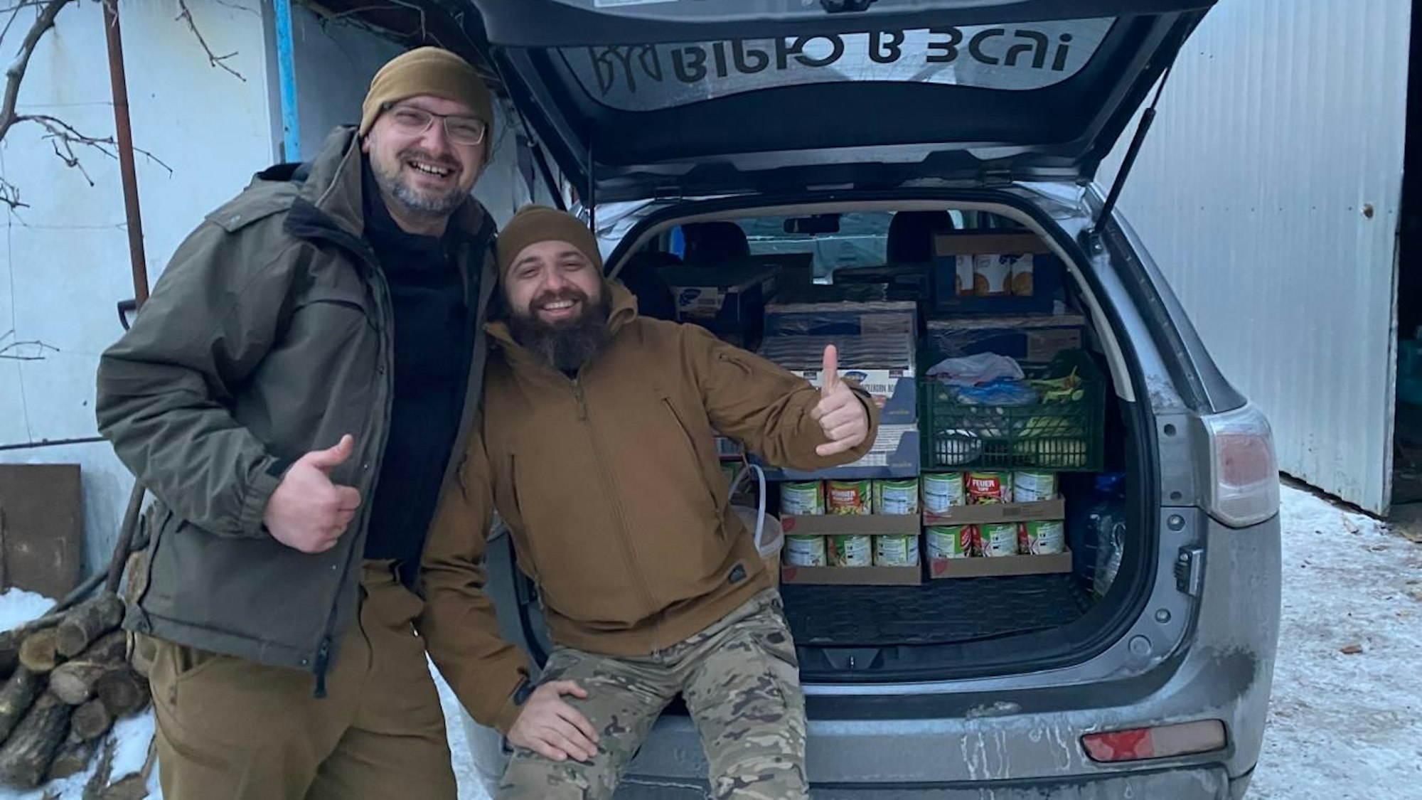 Zwei Männer stehen vor dem offenen Kofferraum eines Autos, in de sich palettenweise Lebensmittel wie Konserven befinden.
