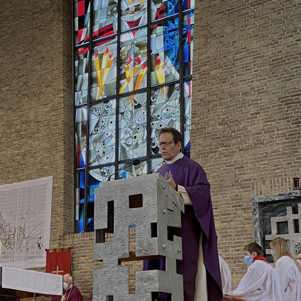 Der Pfarrer trägt eine violette Robe und steht an einem Rednerpult. Im Hintergrund ein Kirchenfenster.