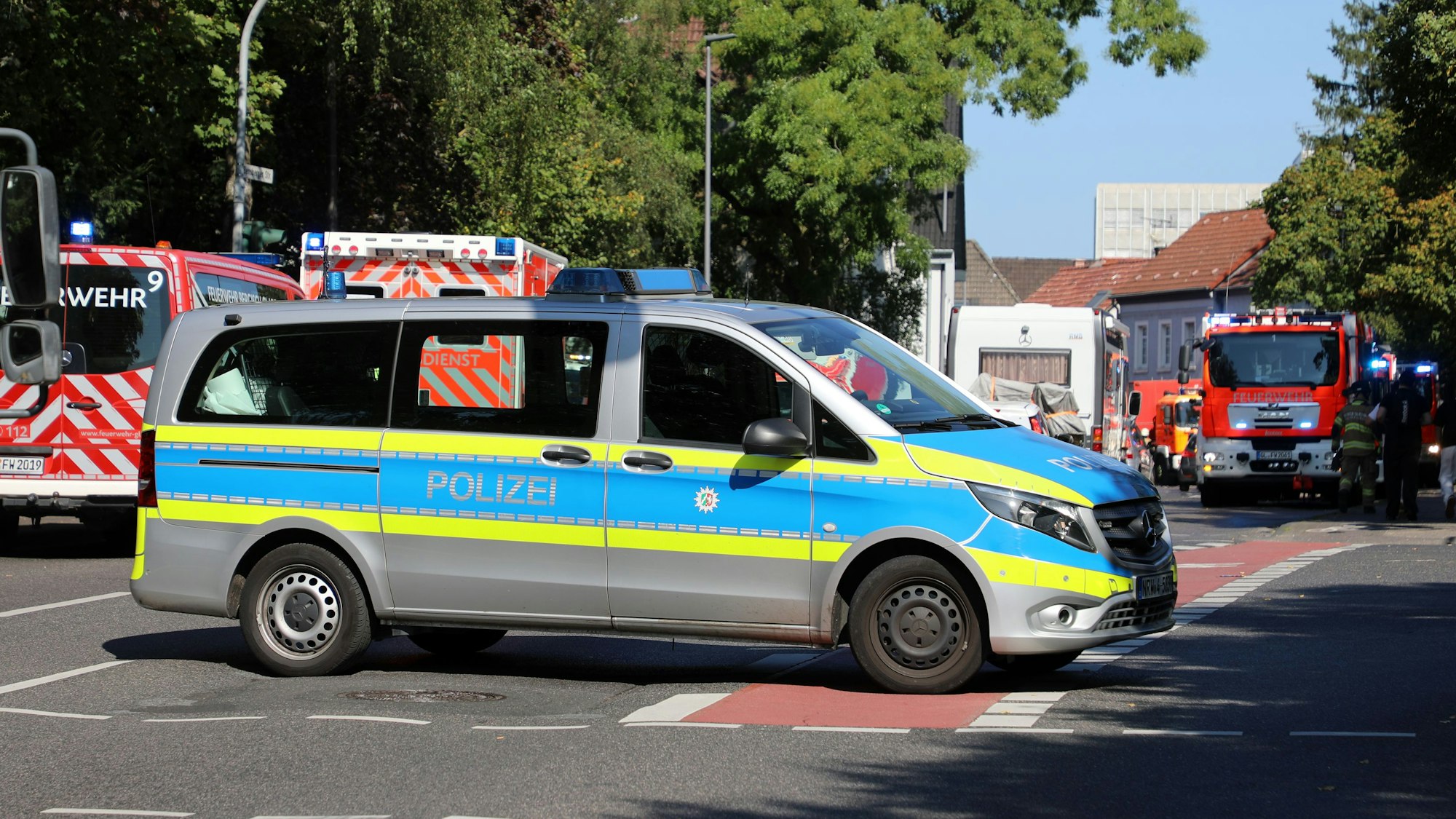 Ein Polizeifahrzeug steht quer auf der Bensberger Straße in Bergisch Gladbach-Heidkamp. Im Hintergrund sind Einsatzfahrzeuge von Feuerwehr und Rettungsdienst zu sehen.