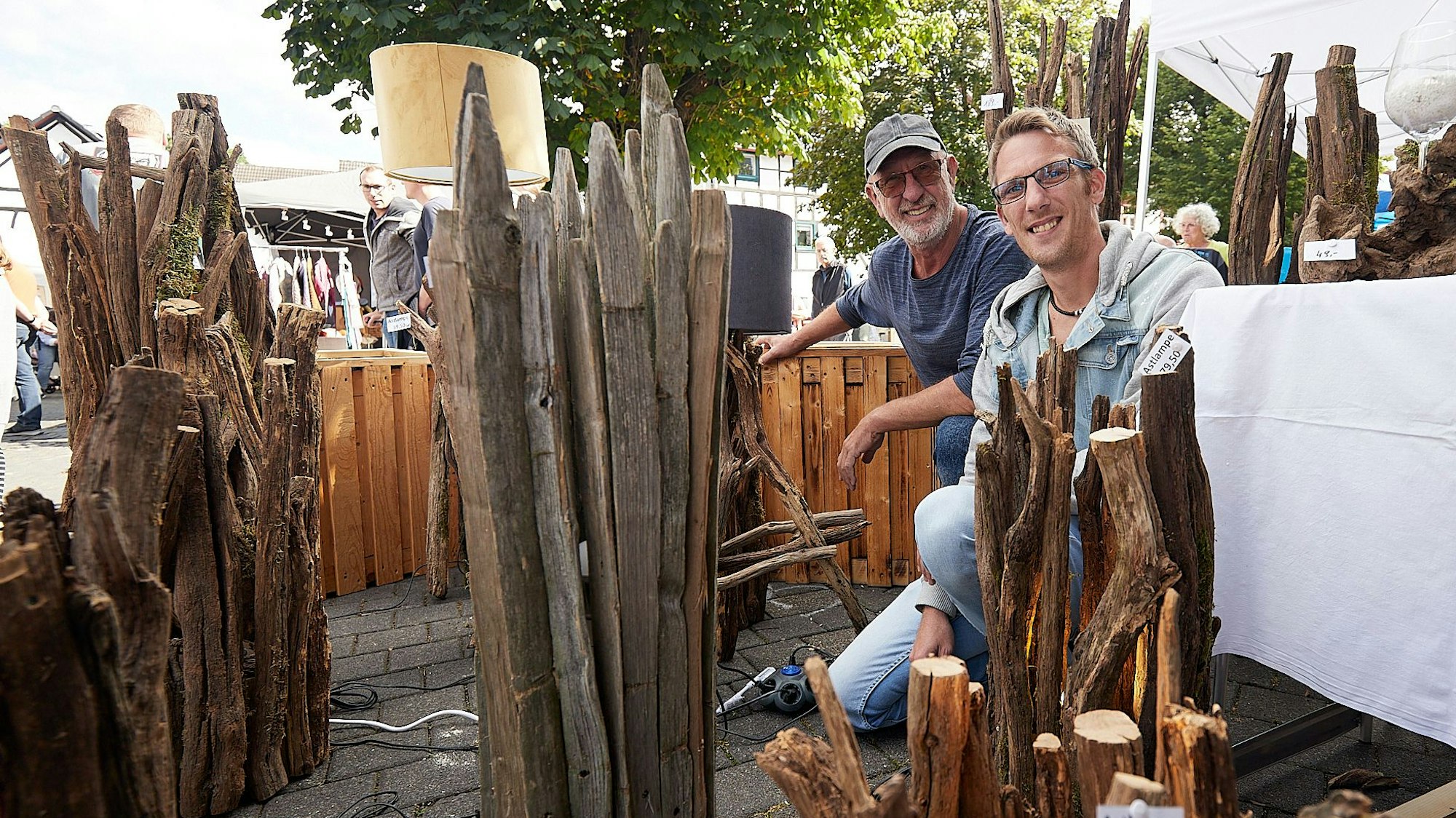 Zwei Männer, Peter (l.) und Kevin Schiller, sind auf einem Markt zwischen den von ihnen angefertigten Lampen aus Ästen zu sehen.