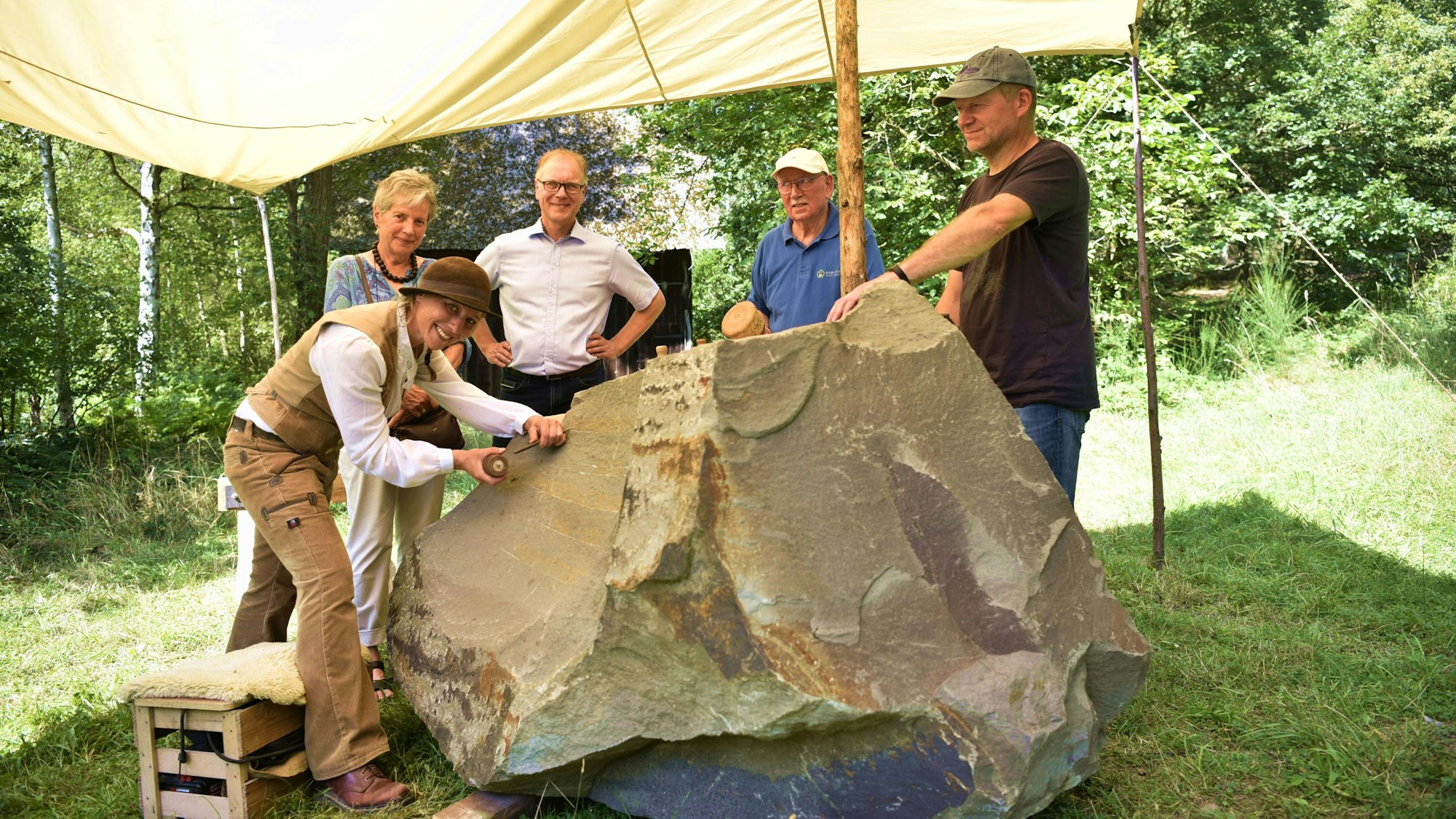 Steinmetzin Ulrike Glaubitz arbeitet in ihrer beigen Handwerks-Kluft unter einem Sonnensegel an dem Gedenkstein für Prof. Dr. Wolfgang Schumacher. Um den Stein herum stehen (v.l.) Rita Schumacher, Dr. Carsten Vorwig, Ralf Sawatzki und Axel Jakob.