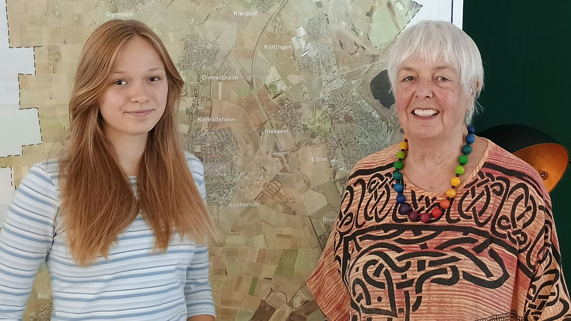 Lena conrady trägt ein weißes Top mit blauen Streifen. Elke Griemens trägt ein rötliches Kleid mit auffälligen Mustern und eine bunte Perlenkette. Hinter den beiden hängt eine große Karte von Erftstadt.