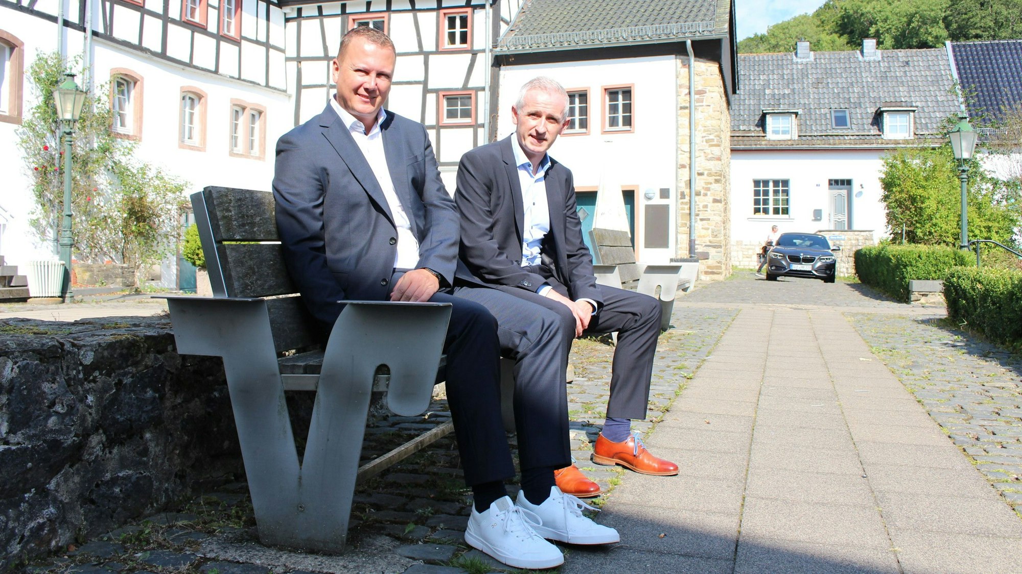 Mark Heiter und Kai Zinken sitzen auf einer Bank. Im Hintergrund sind Blankenheimer Fachwerkhäuser zu sehen.